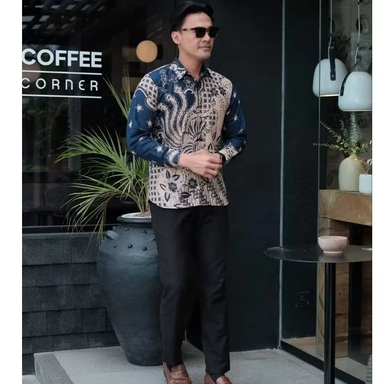 Back view of man wearing premium batik shirt showing intricate navy and cream pattern design with flowing swirls and floral elements on the back panel, standing next to decorative plant pot.
