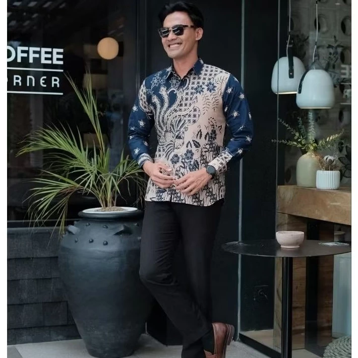 Smiling man in stylish navy blue and cream batik shirt with mixed floral and geometric patterns, brown leather shoes, standing confidently outside coffee corner cafe, modern Indonesian fashion.