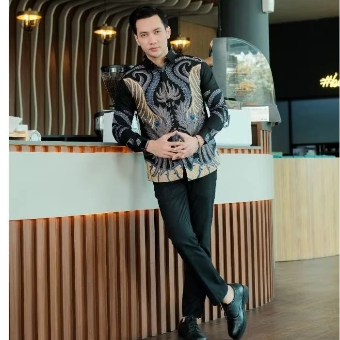 Male model leaning casually against a counter with one hand in his pocket, showcasing a premium black batik shirt with intricate gold wing patterns and blue feather details for a smart casual look.