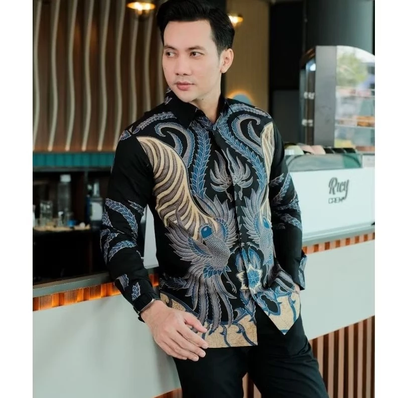 Medium side-angle shot of a man wearing a detailed black batik shirt featuring a large blue and gold bird design, looking away thoughtfully while leaning on a cafe counter.