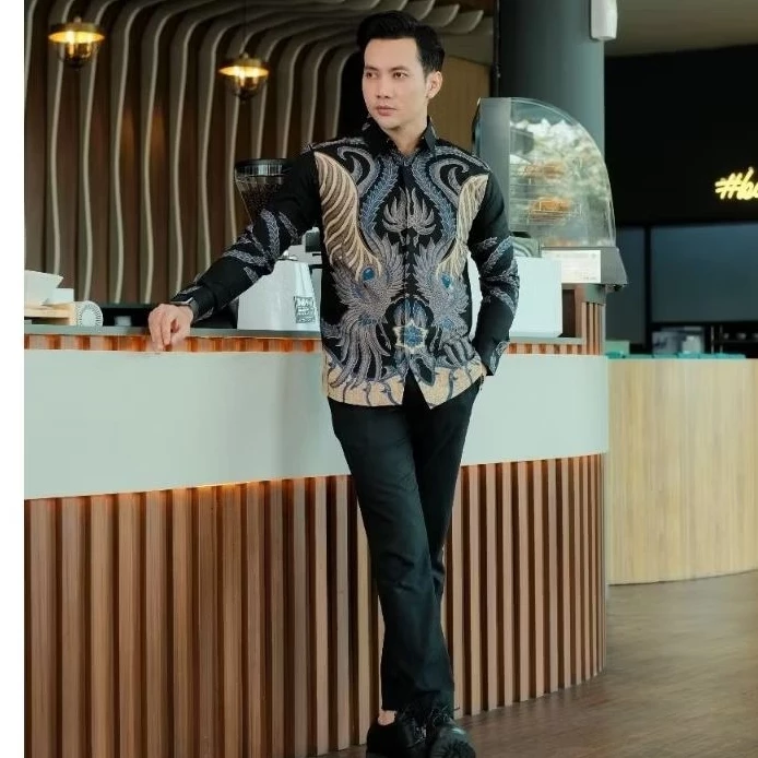 Handsome man in a stylish black and gold batik shirt leaning against a white counter with legs crossed and hands clasped, modeling modern Indonesian executive formal wear.